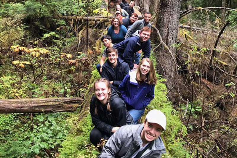 A group of students pose lined up on a mossy log in the forest.